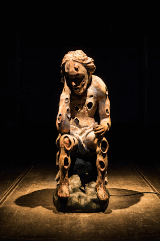 Close-up of the brown sculpted figure with a warm light spotlight illuminating it from above. The figure is sitting down with their hand holding their head, in a reflexive pose. They have circular carvings all over their body. This is part of an art installation in Tramway, Glasgow International 2018.