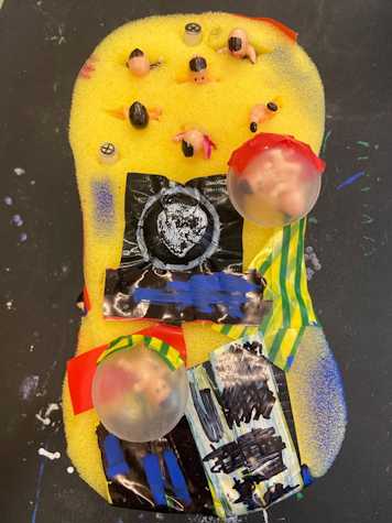 A large yellow cleaning sponge that has been decorated with different coloured paints, assorted types of tape, and mini human figurines.