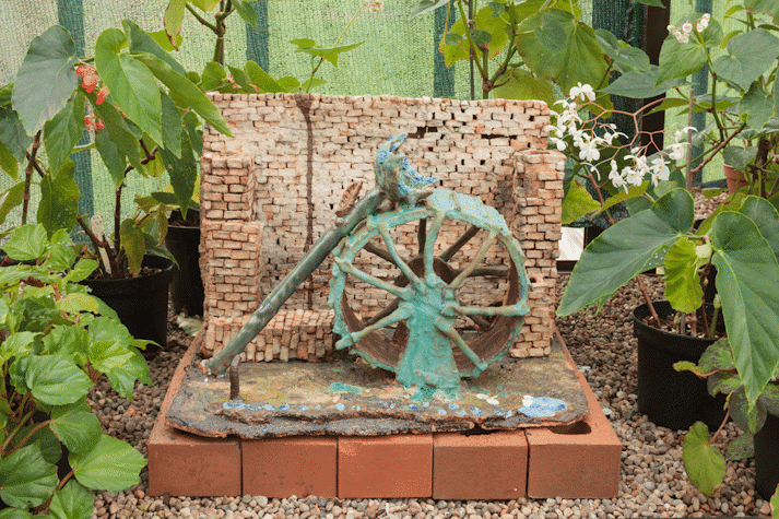 In a greenhouse, between plants, a small display of a ceramic green wheel with a flower on top of it, on top of a base of bricks.
