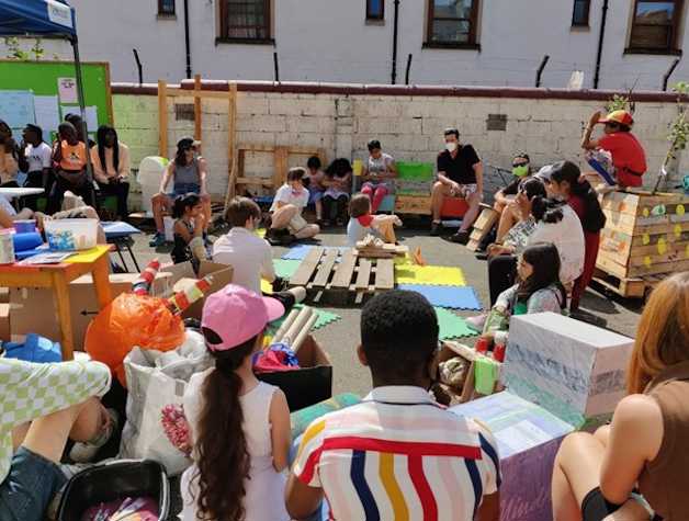 A photograph featuring a large group of people sitting in an outdoor space in front of a building. The space is also filled with gardening and creative supplies.