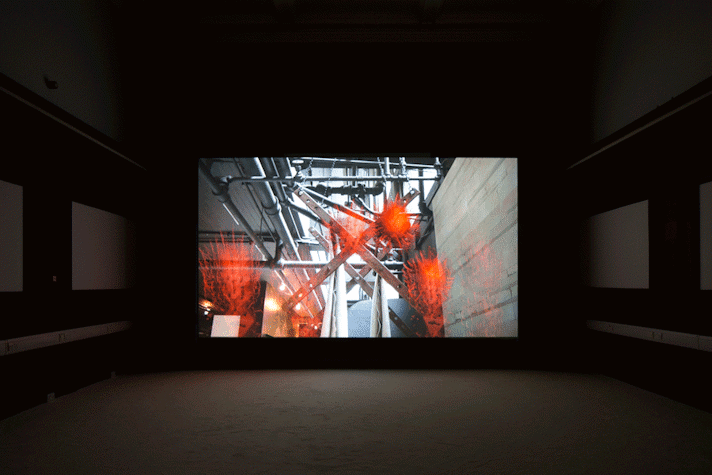 In a dark room, there is a large projector screen placed in the centre. This screen shows the image of a wall on the side and pipes spread out throughout the area. There are some orange translucent work like objects, made of hay like material, placed on various parts of the image. The walls on the sides of the screen have some screens hanging on them, but they are barely visible due to the darkness.