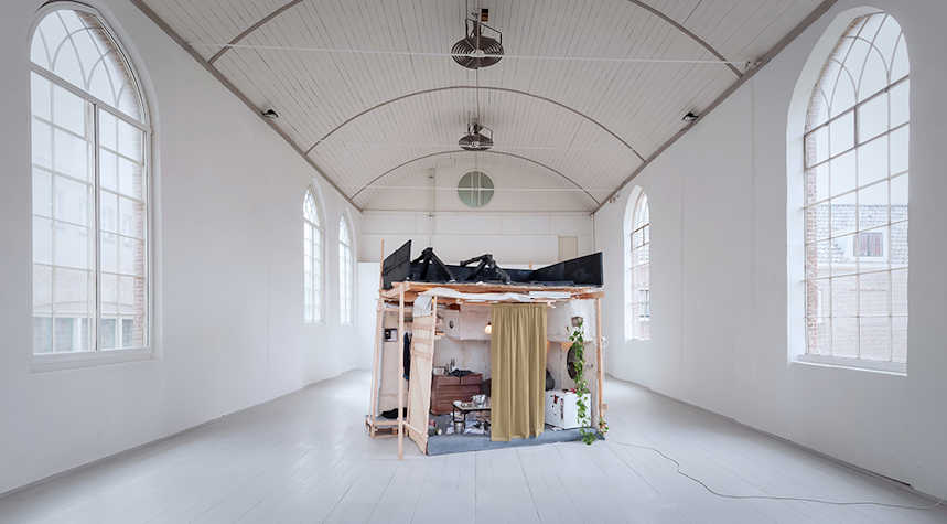 A room with white walls and floors and large windows with natural light streaming in. At the centre of the room is an art installation of a home space featuring a storage furniture and a partially obscured armchair.