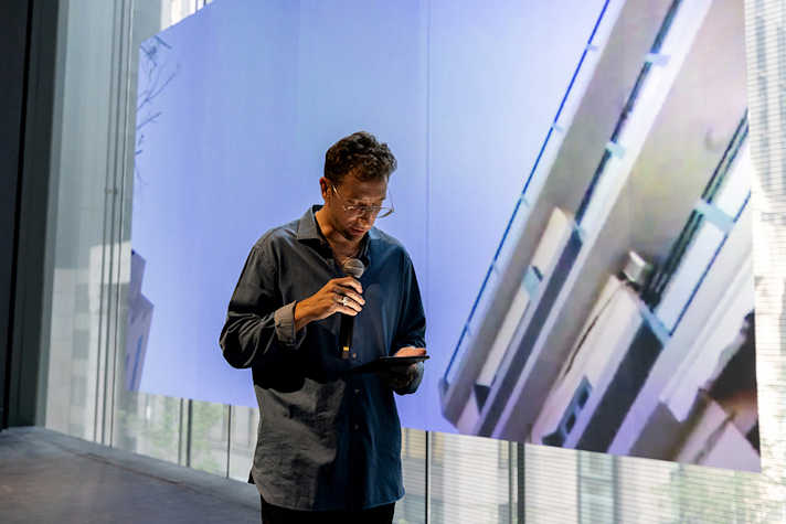A man stands on a stage area. He holds a microphone to his face with his right hand and is looking down at a tablet held in his left hand