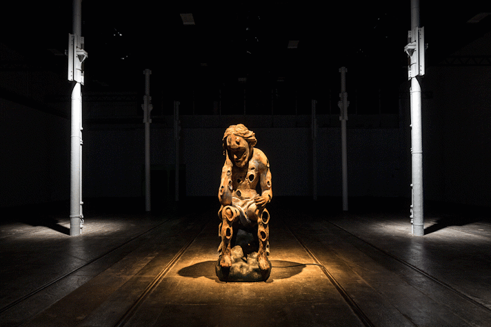 There is a dimly lit room with four thin metal grey columns and wooden floors. We can see a brown sculpted figure in the middle with a warm light spotlight illuminating it from above. The figure is sitting down with their hand holding their head, in a reflexive pose. They have circular carvings all over their body. This is part of an art installation in Tramway, Glasgow International 2018.