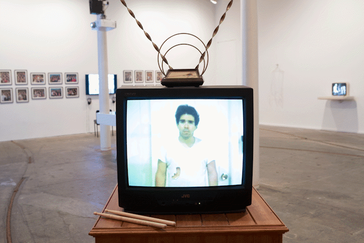 A small television monitor is placed on a table slightly bigger than the monitor. There are a pair of drumsticks sitting next to the monitor and antennas on top of it. The screen of the monitor shows a man, looking sad, in a torn white t-shirt. The wall behind the monitor has rows two rows of framed pictures with a television screen right in the middle of them. There is a chari in front of the screen. On the right side wall there is another small television monitor, its picture is blurry.