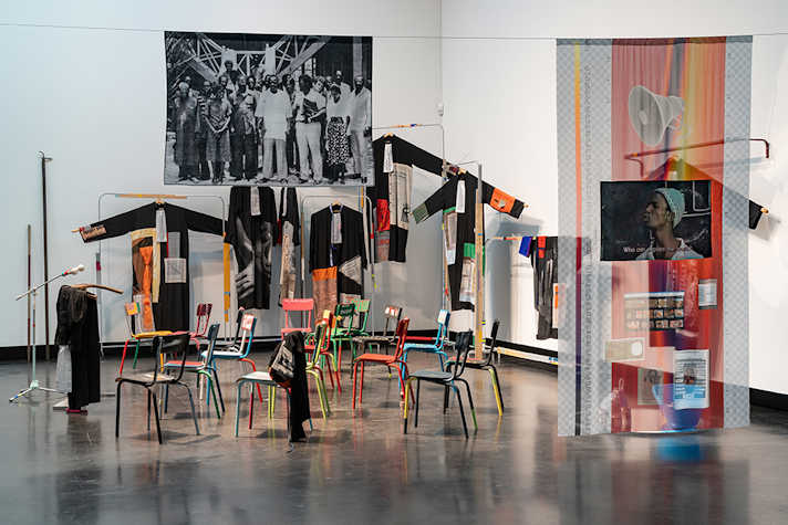 A room with white walls, the walls are covered with hanging artwork and coats, and in the centre are a selection of brightly coloured chairs with a lectern and microphone in front.