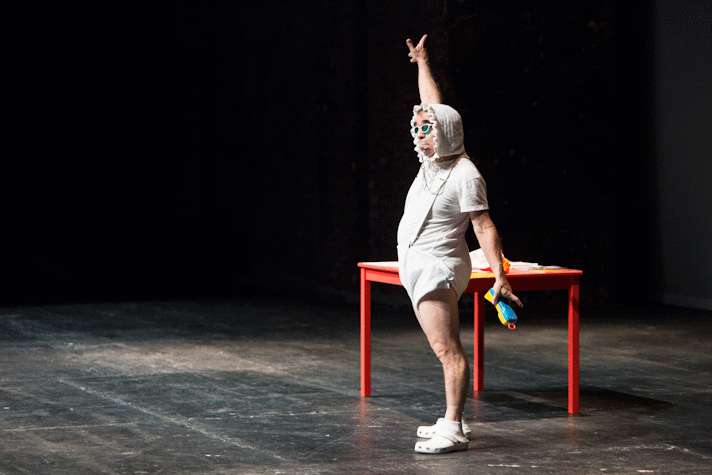 In a dark room there is a spotlight on a man wearing a baby attire. He is wearing a diaper, a vest, bib, hat, crocs and sunglasses. He has a pacifier in his mouth and a toy telephone in one hand. His other hand is pointing towards something in the distance above his head.l There is a table behind him. The table has stacks of papers.