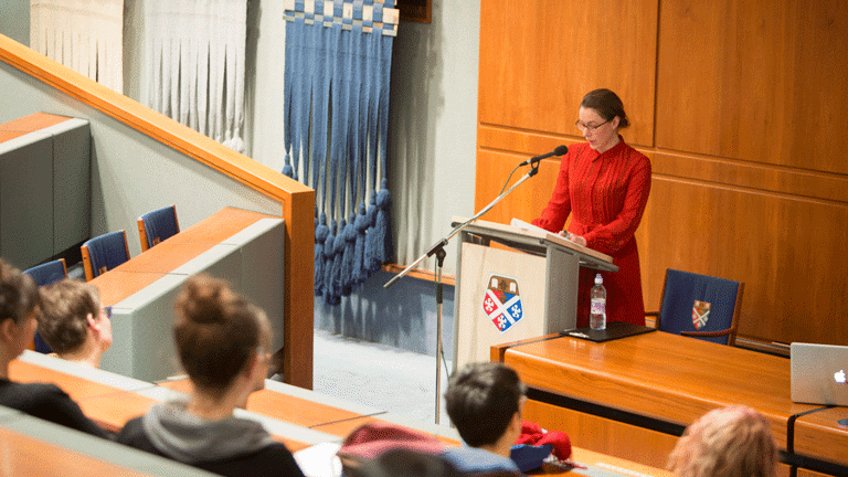 In a lecture theatre, a woman is giving a speech, through a microphone, standing behind a podium. There are many people sitting in front of her on benches, in the lecture theatre.