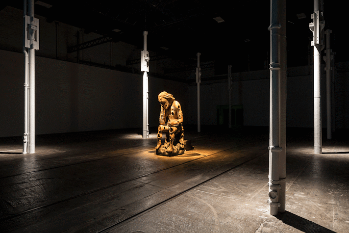Diagonally view of a dimly lit room with four thin metal grey columns and wooden floors. We can see a brown sculpted figure in the middle with a warm light spotlight illuminating it from above. The figure is sitting down with their hand holding their head, in a reflexive pose. They have circular carvings all over their body. This is part of an art installation in Tramway, Glasgow International 2018.