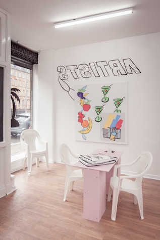 The room has an tall open window on the left side, the set up is that of a single table nail salon. There is a small table with two chairs facing each other placed in the centre of the space. There is a painting on the wall, it has fruits, cocktail glasses and paint buckets painted on it. On the wall, the words "ARTISTS" is written as a mirror image, with a half a paintbrush peeking from under the hung painting.