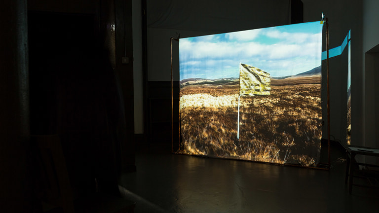 In a dark room a film is projected onto a screen. The project image shows a flag made from a gold emergency blanket, flying in an open field with hills in the far distance and a blue sky