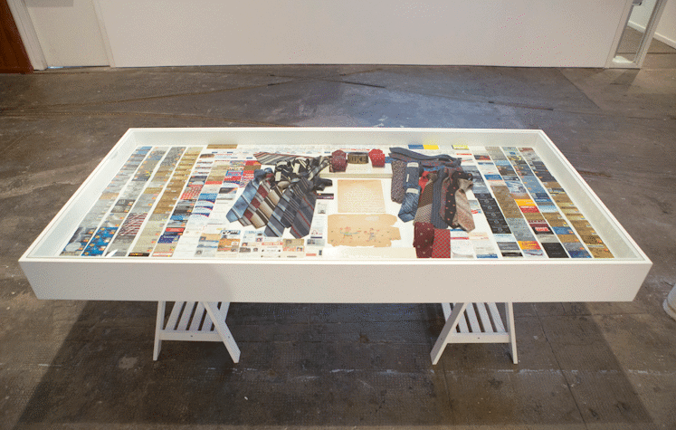 An archival table is placed on the ground, which has spots of paint and dirt on it. The table showcases a spread of credit or debt cards on the sides, surrounding a collection of ties. The ties are made to surround some archival papers and identity cards. The lengths of the table have rows of business cards. There is a thick block placed above the sheets, it holds two pairs of rolled ties and in the middle there is a rolled up belt which reads "MIKE".