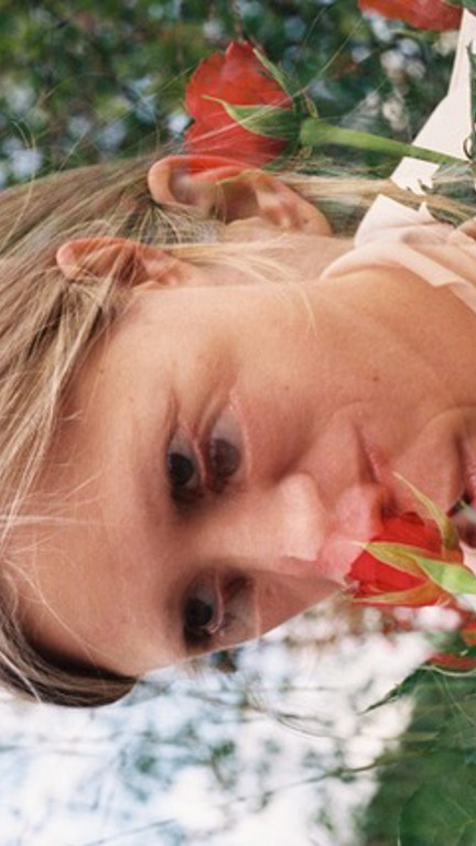 A blurred, overlapping image of a person with blonde hair which is tied back, they have flowers near their mouth and head.