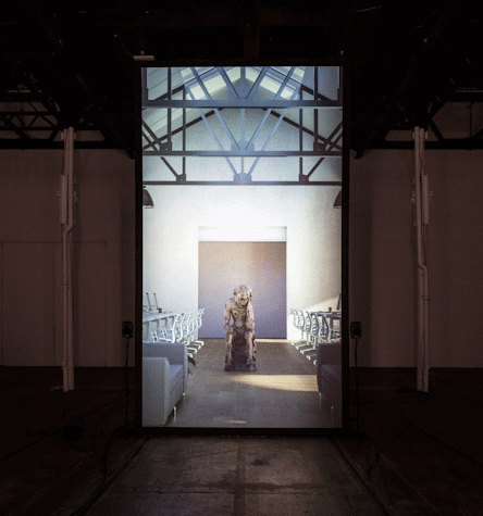 Big screen with a picture of the brown sculpted figure with a warm light spotlight illuminating it from above. The figure is sitting down with their hand holding their head, in a reflexive pose. They have circular carvings all over their body. This is part of an art installation in Tramway, Glasgow International 2018.