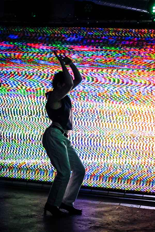 A performer in a black leather top and cream trousers is silhouetted mid-movement against a large screen of glitching, multicoloured static, arms raised above their head.