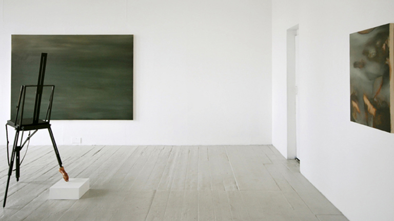 two dark grey green blurred paintings on adjacent walls. in the middle of the room is a black easel,