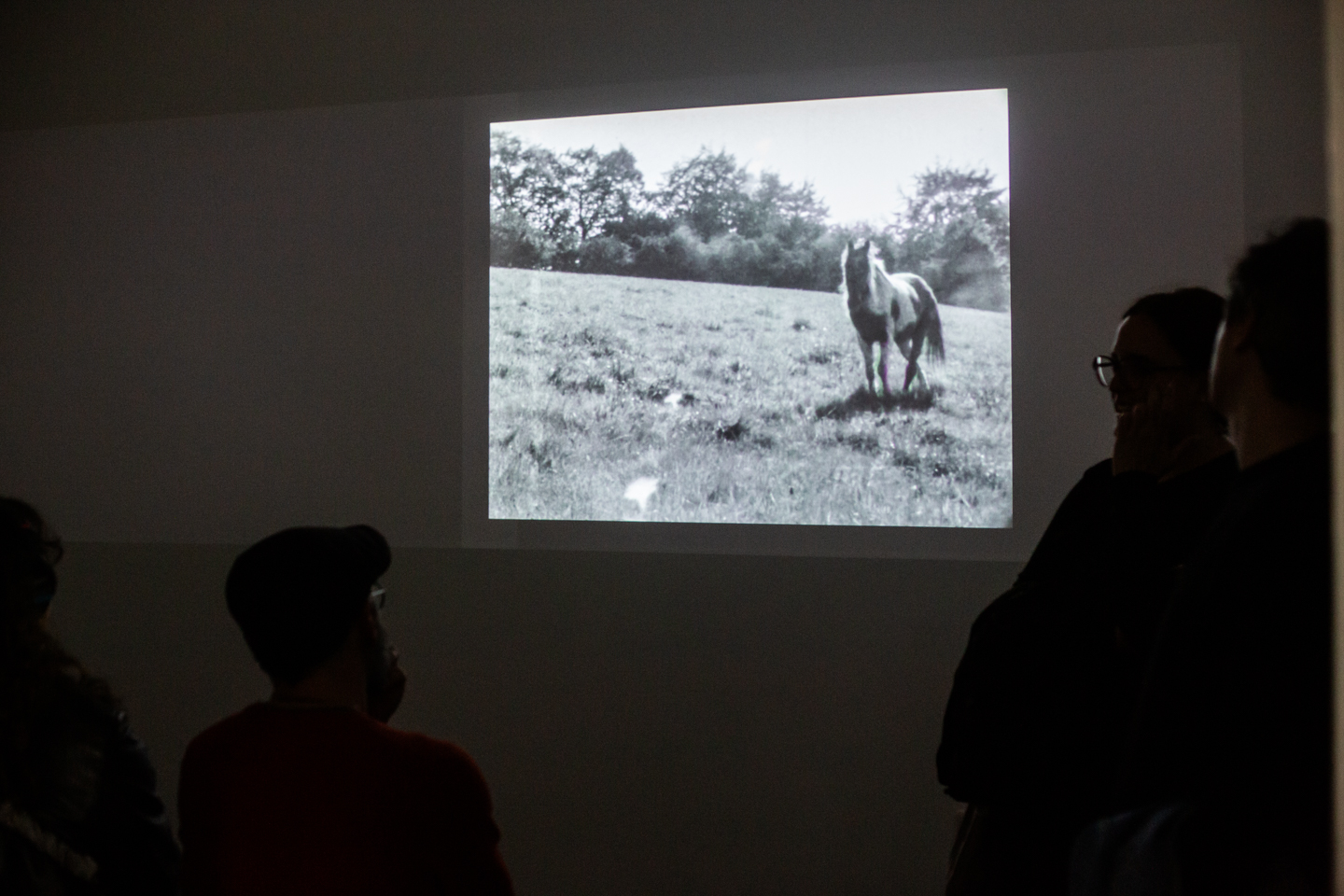 Visitors silhouetted on a bench watch two adjacent film projections, one showing dark figures on a sloped rooftop and the other an arched monument against a pale sky.