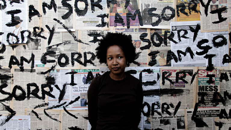 A woman with a black top stands against a wall of collaged newspapers, that says "I am sorry".