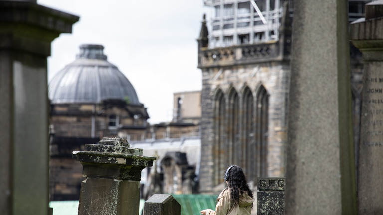 A person wearing headphones and a beige jacket walks through Glasgow Necropolis, gravestones in the foreground and the Gothic tower of Glasgow Cathedral visible behind scaffolding in the background.