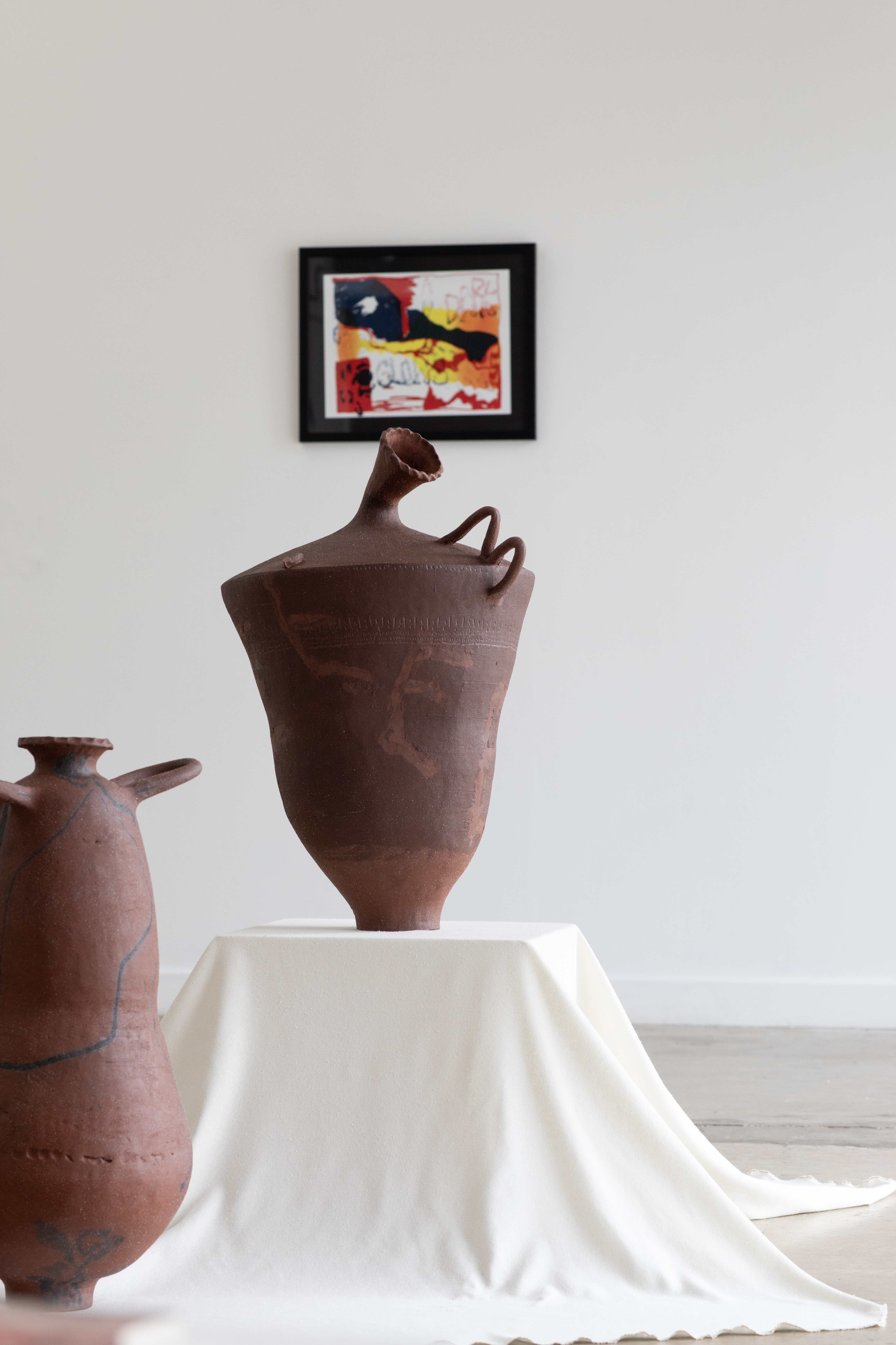 A tall, urn-shaped terracotta ceramic sculpture with a narrow spouted neck and looping handle stands on a white draped plinth, beside another handled vessel, with a small framed abstract painting on the wall behind.