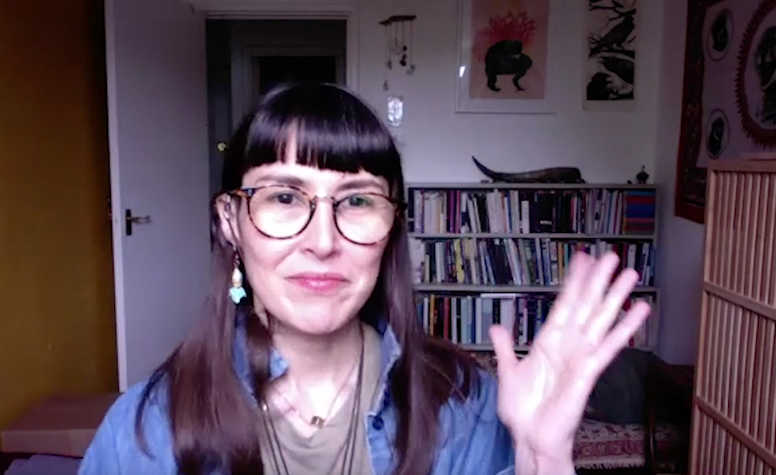 A photo of a woman with brown hair and glasses who is sitting with her right hand raised in a wave. The room in the background contains a bookcase and photographs on a white wall.