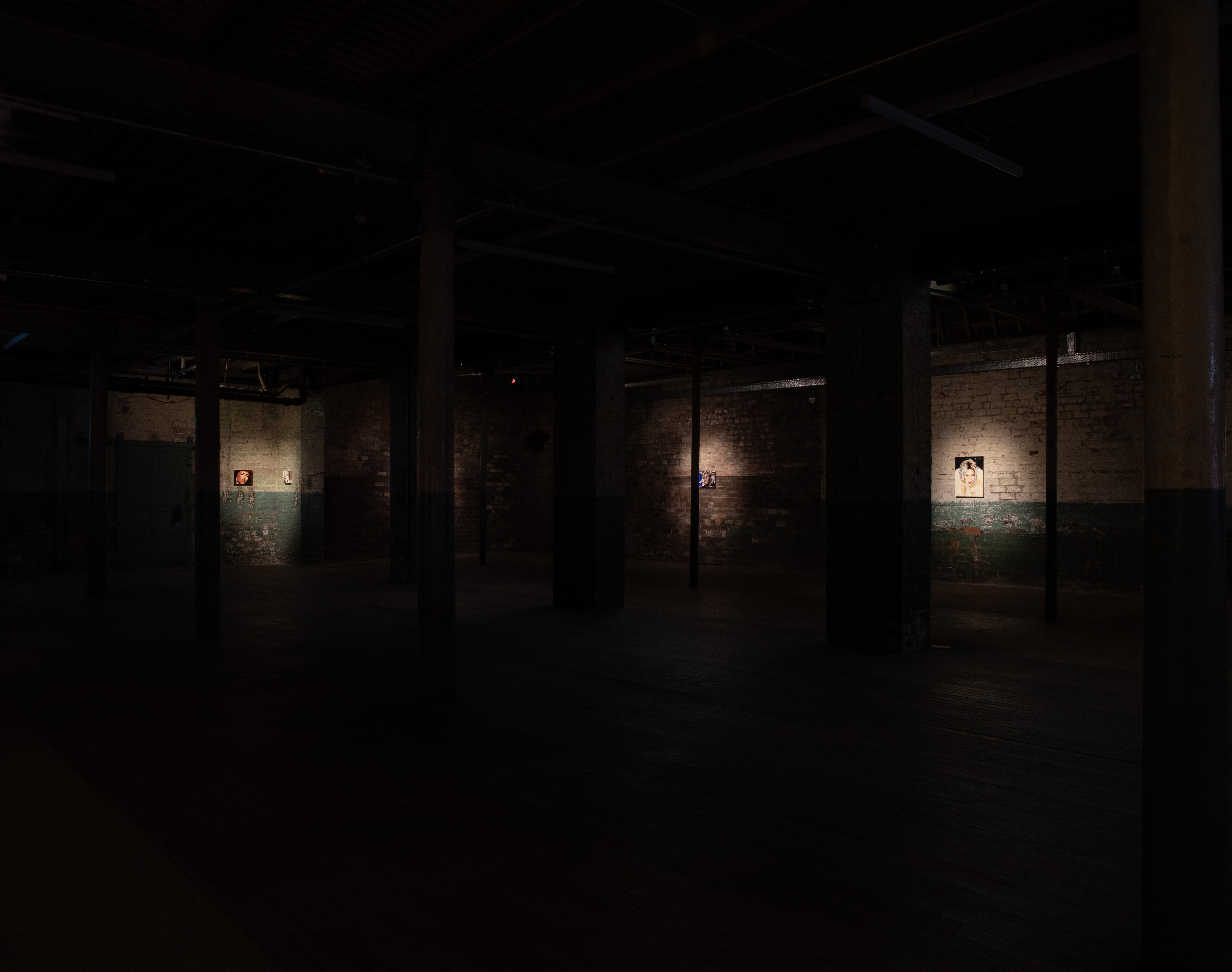 Wide, dimly lit gallery space with rows of columns and exposed brick walls. Small spotlit portrait paintings are visible on three separate walls in the distance, sparsely placed across the dark room.