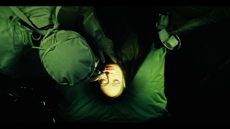 Colour film still depicting a surgical scene from above. A patient lies on an operating table with their face illuminated against darkness while medical professionals in green surival gowns lean over them. Surgical instruments are visible, including a reflective instrument or mirror. The dramatic lighting and palette of surgical greens and warm skin tones emphasise horror-film qualities and themes of bodily transformation.