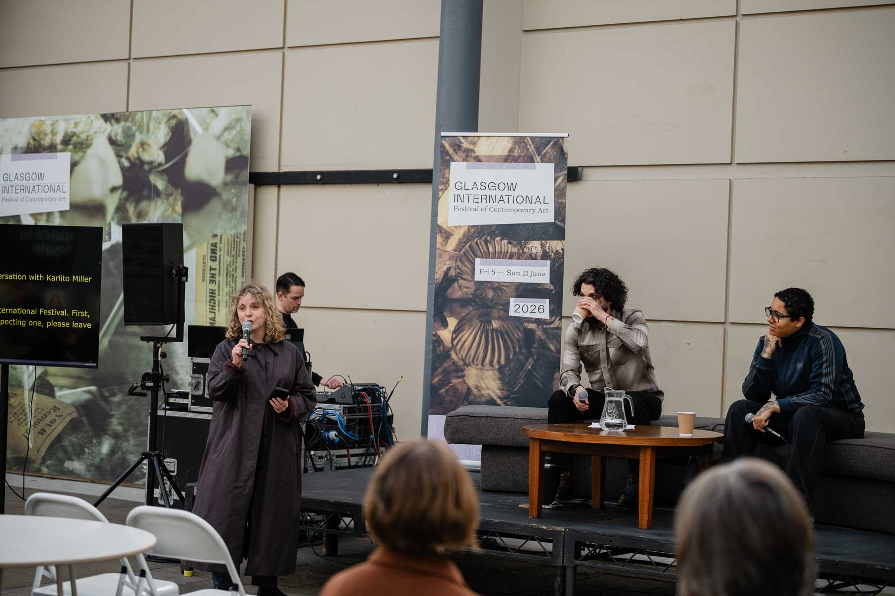 A woman stands introducing an event into a microphone, with a screen behind her reading "conversation with Karlito Miller." Two men sit talking on a bench-style seat further along, with Glasgow International banners either side of them.