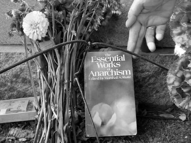 black and white image. there is a book called the essential works of anarchism edited by Marshall s shatz against a brick wall. someone's hand reaches towards the book. a bunch of flowers lean against the wall next to the book.