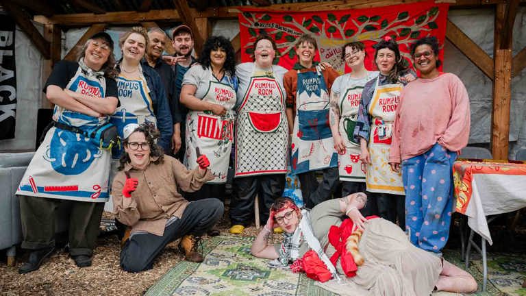 Group portrait of Rumpus Room volunteers wearing matching printed aprons under a wooden shelter, laughing together in front of a red banner appliquéd with tree branches, with two people play-acting on a patterned rug in the foreground.
