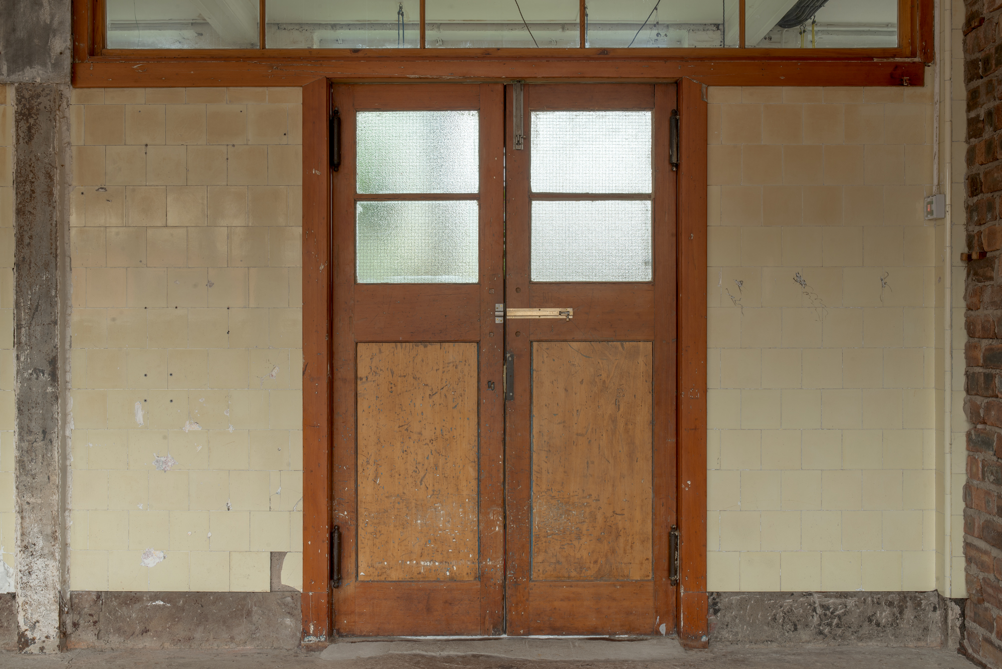 A weathered wooden double door with frosted glass panels set into a tiled corridor wall, original fittings and peeling paint visible.