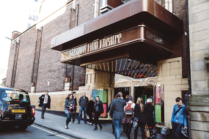 The outside of a theatre can be seen. It has the words "Glasgow Film Theatre" and "Cafe Custom" attached on the entrance. There is a crowd of people entering the building. A taxi is standing outside the theatre.