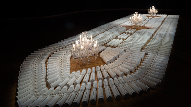 Plaster cast replica tamtam drums arranged in the shape of the hold of a ship against a dark backgrounds, three chandeliers placed above the work horizontally