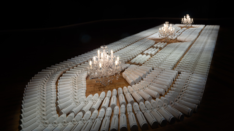 Plaster cast replica tamtam drums arranged in the shape of the hold of a ship against a dark backgrounds, three chandeliers placed above the work horizontally