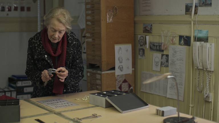 Film still showing an older woman measuring something carefully. In the background are various items that suggest this is an archive 