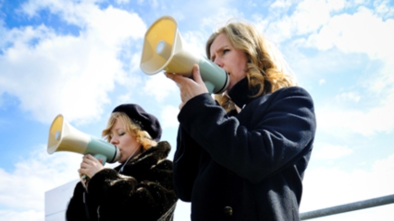 two women speaking into megaphones standing outside behind a yellow barrier