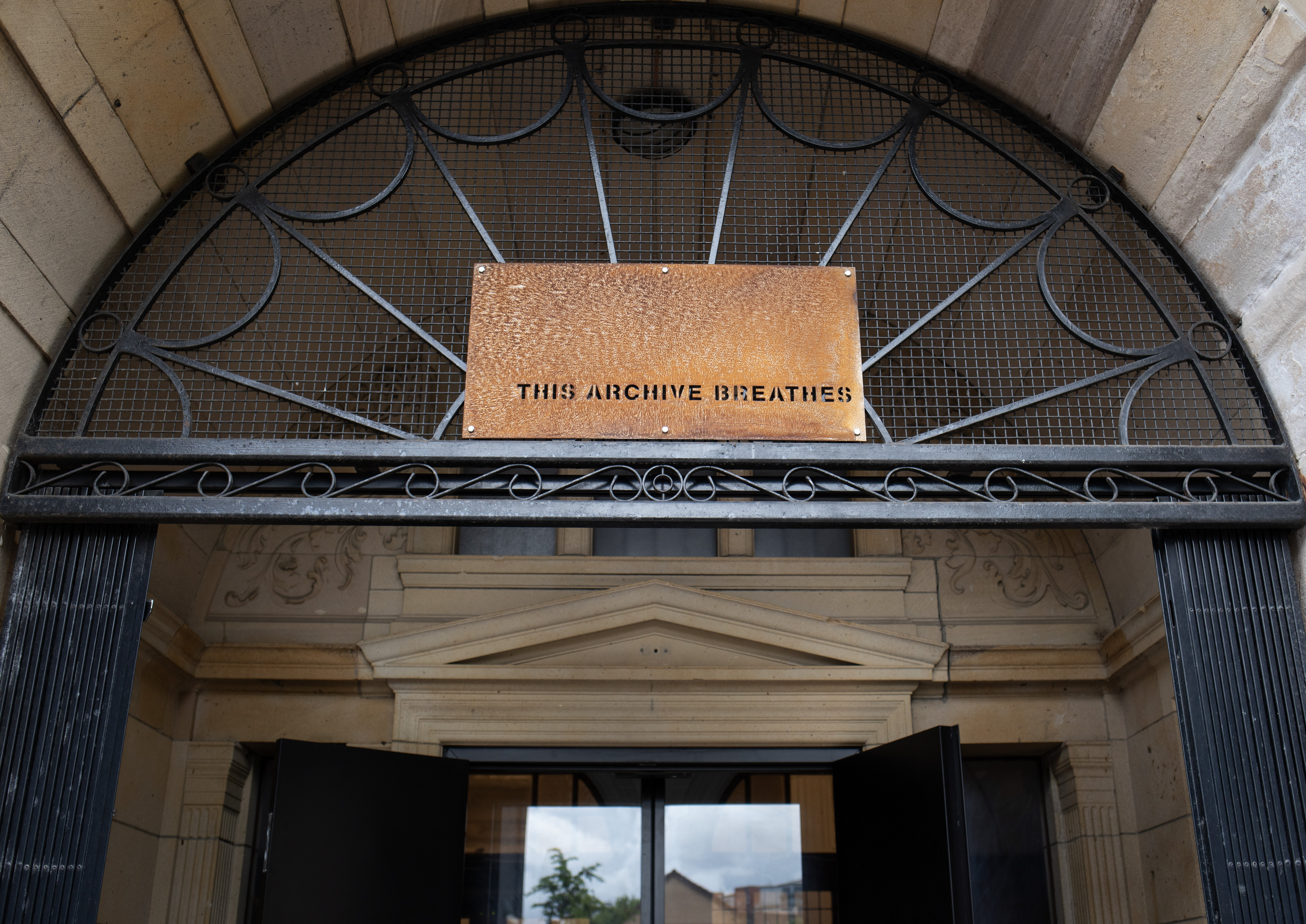 A copper plaque reading "This Archive Breathes" mounted within an ornate wrought-iron fanlight above a building's entrance.