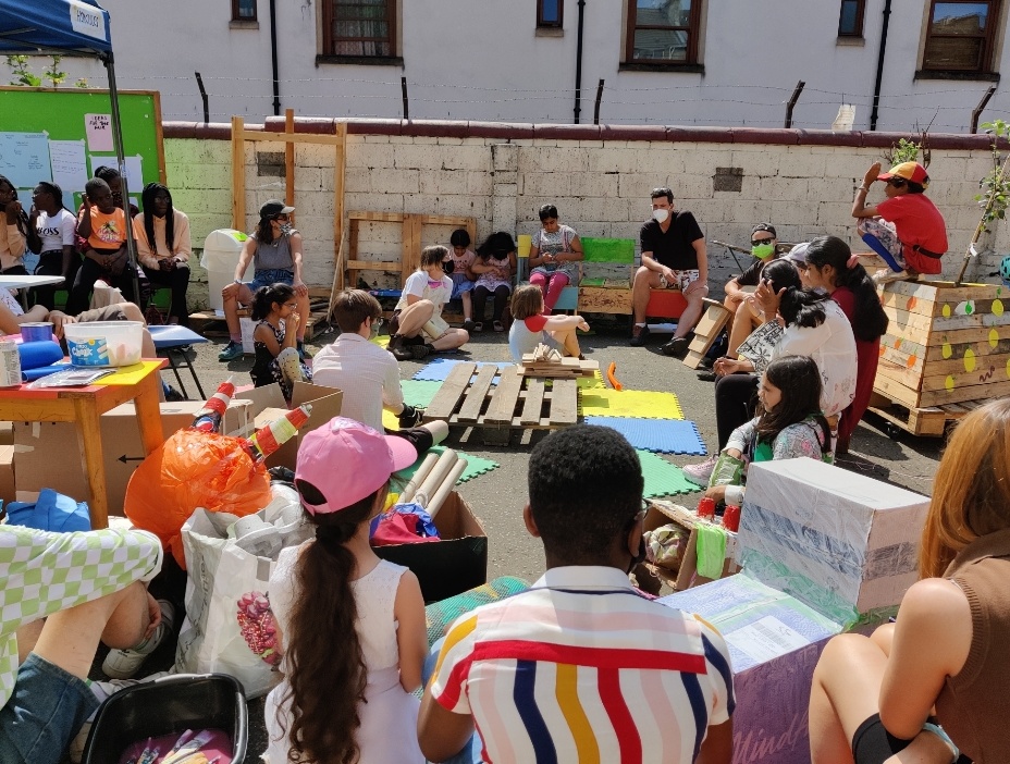Yard School. Photograph of children and adults sitting together outside in the sunny yard at Rumpus Room. Wooden crates, painted cardboard boxes, and other materials are scattered around them. 