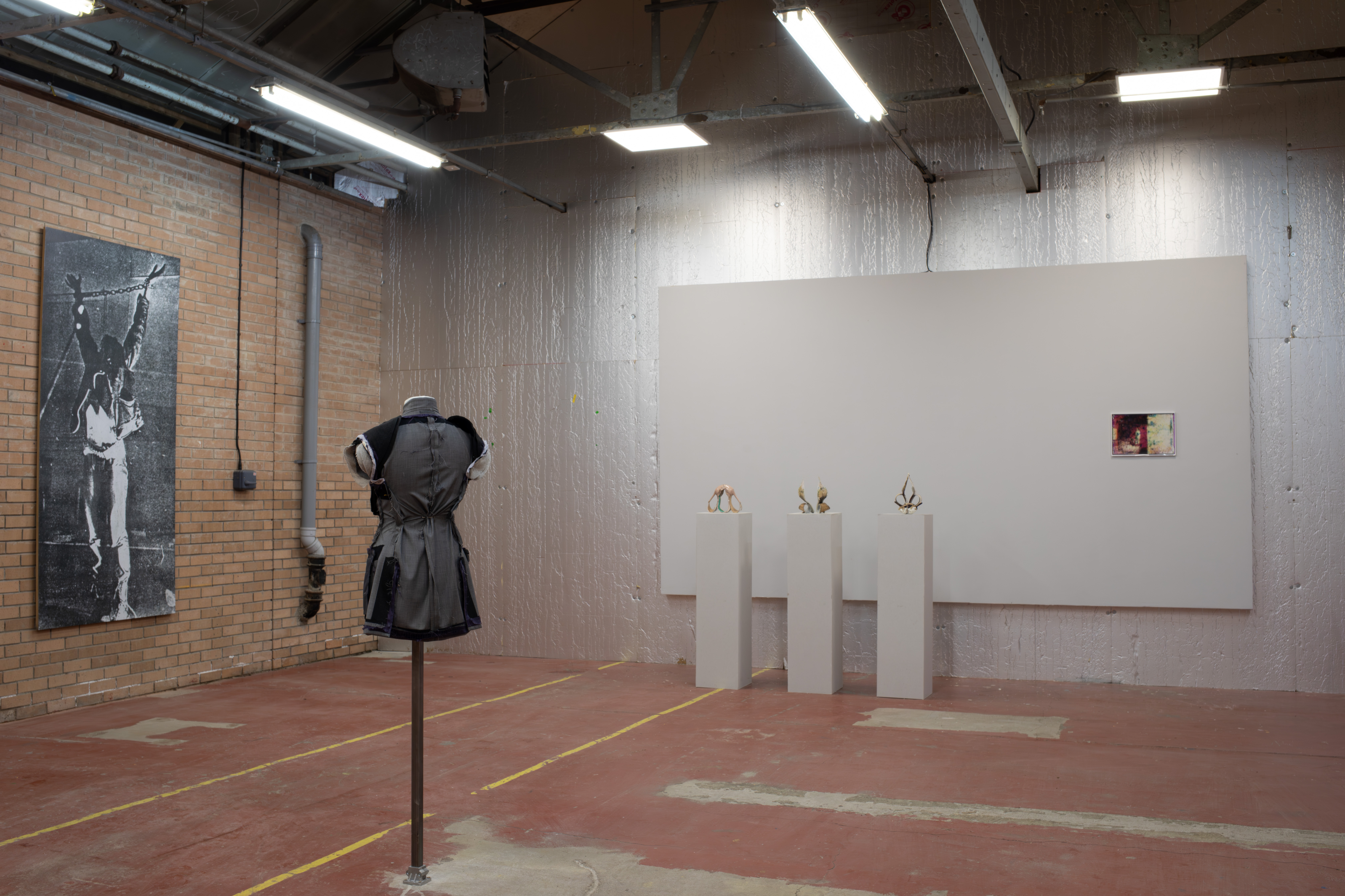 An industrial gallery space showing a black and white photograph of raised, chained hands on brick, a headless grey garment on a stand, small jewellery pieces on plinths, and a framed photo on a silver wall.