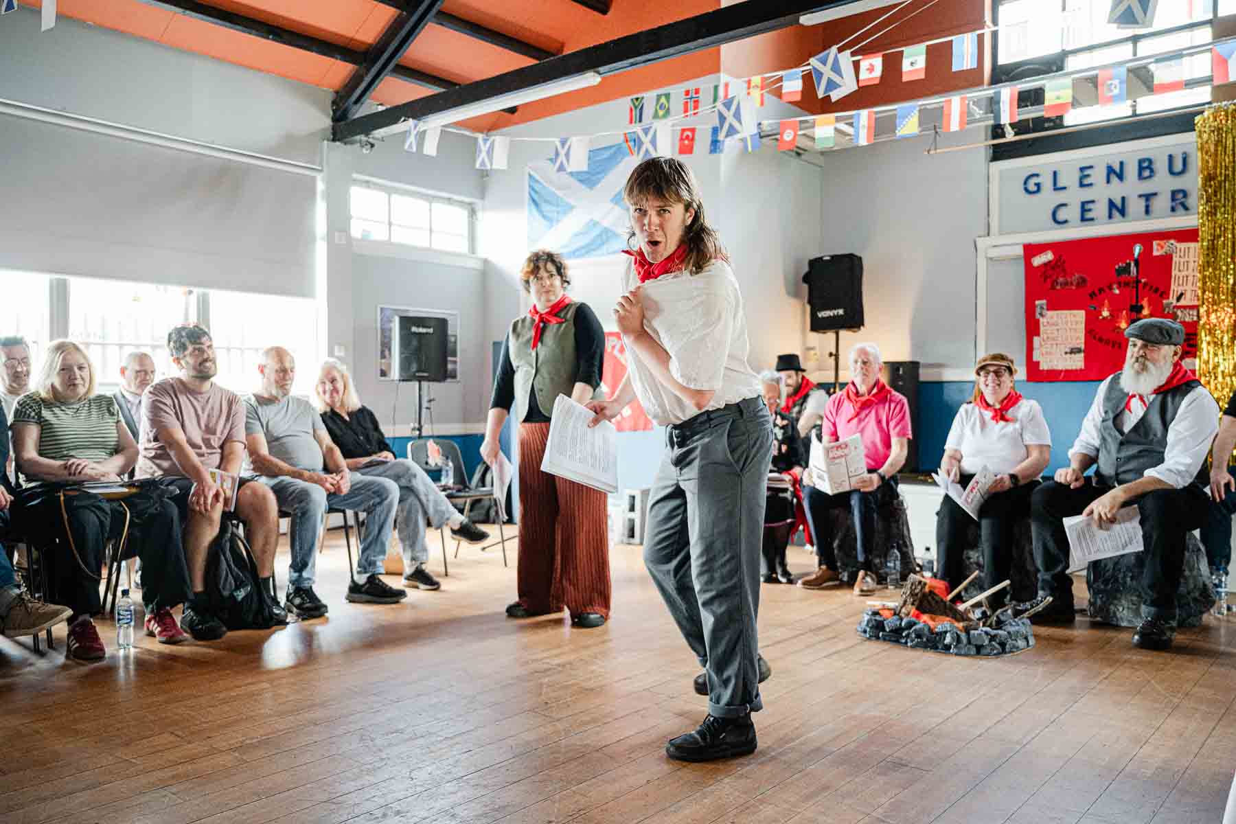 A performer in a white shirt and red neckerchief strikes an animated pose mid-scene, with seated audience members and cast in the background, flags and bunting overhead.