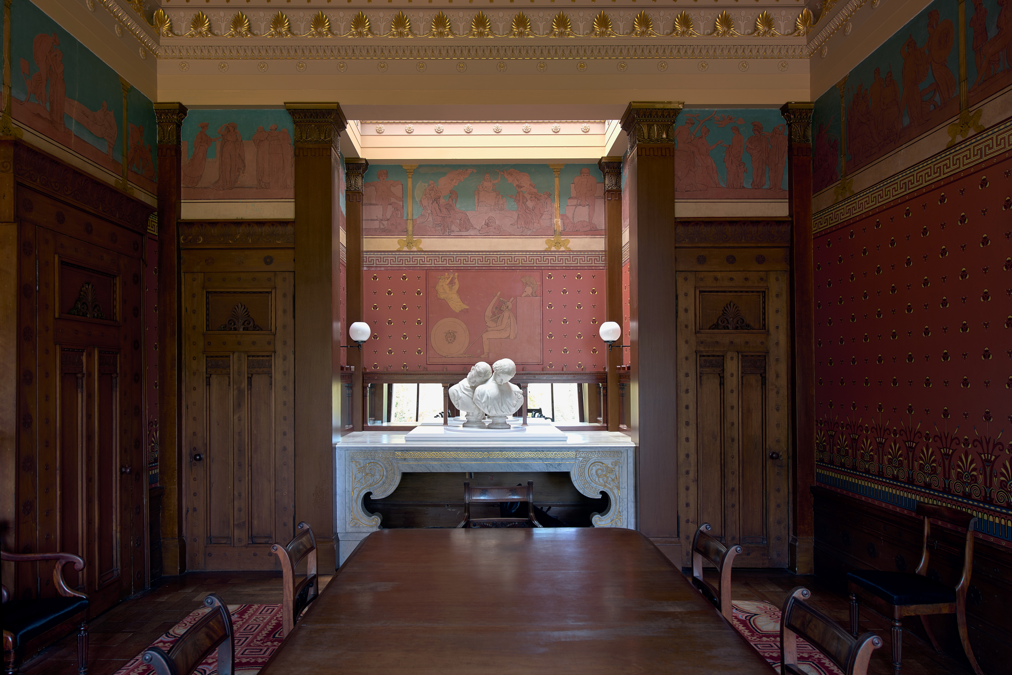 A grand painted room at Holmwood House, its walls decorated with classical friezes in red, teal and gold, with a marble fireplace and a white marble double bust as part of Jon Owen's installation.