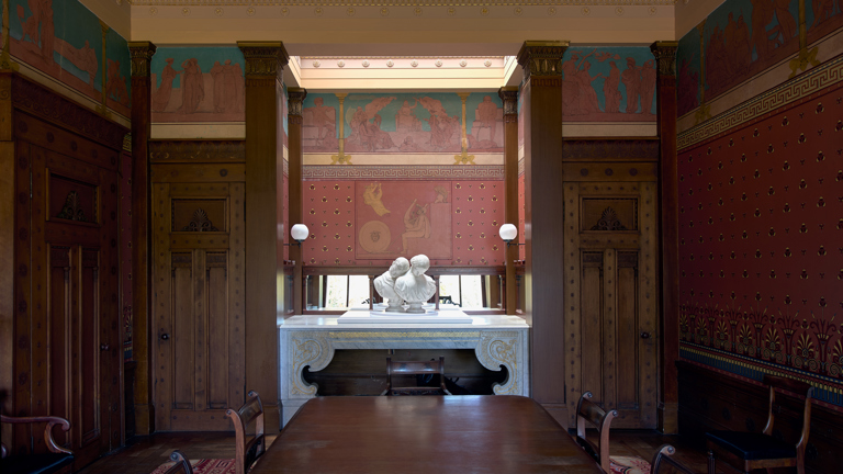 A grand painted room at Holmwood House, its walls decorated with classical friezes in red, teal and gold, with a marble fireplace and a white marble double bust as part of Jon Owen's installation.