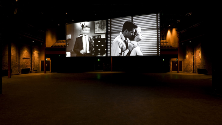 two projection screens hanging side by side in tramway 1. on the left is a man standing in a suit. on the right is a person stood behind another person with their hands on their shoulders in embrace.