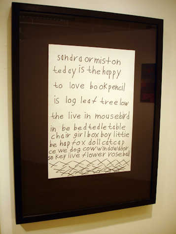 white piece of paper in a black frame. the paper reads: "tedey is the happy to love book pencil is log leaf tree low the live in mousebird in be bed tedle table chair girl box boy littie be hap fox doll cat cap ce we dog cow window door so key live flower rose ball xxxxxxxxx