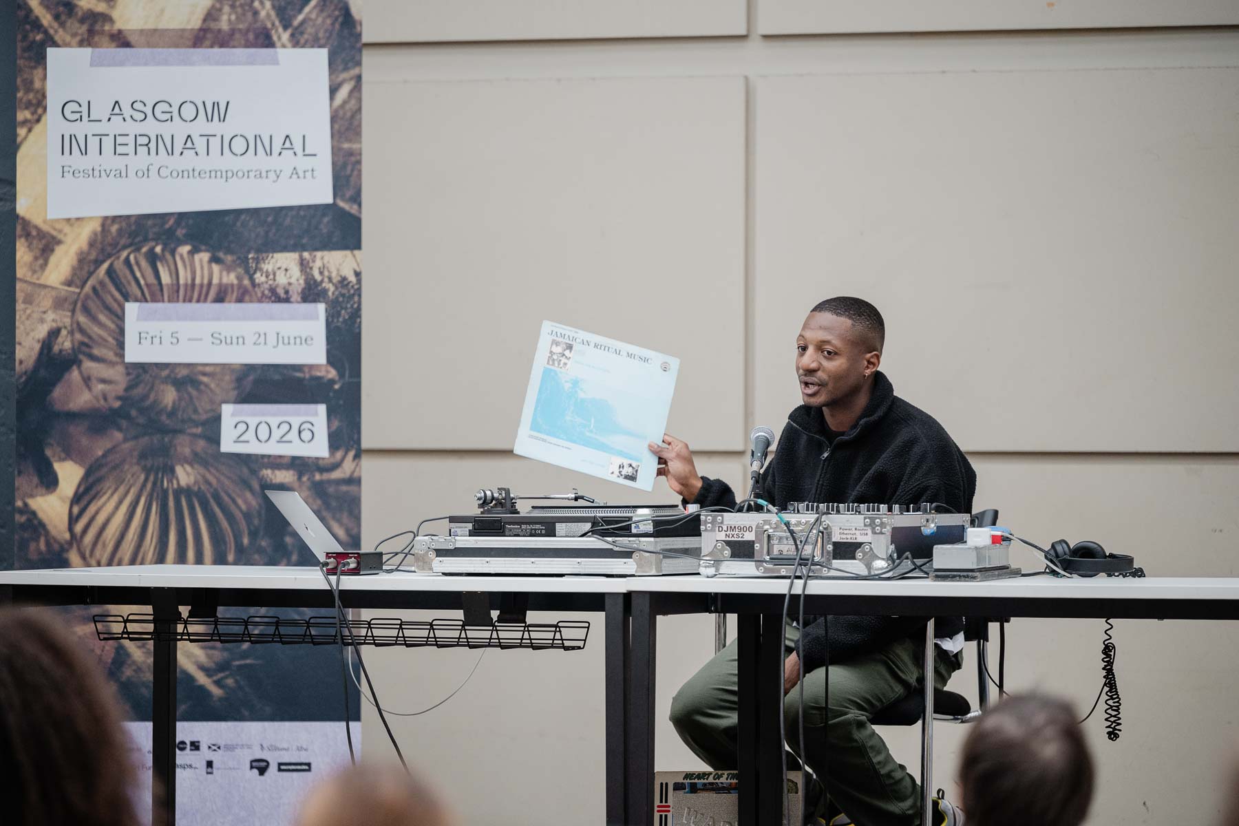 Ashley Holmes sits at a table set up with DJ equipment, including turntables and a mixer, holding up a record sleeve reading "Jamaican Ritual Music" and speaking into a microphone. A Glasgow International festival banner stands behind him, listing the festival dates as 5 to 21 June.
