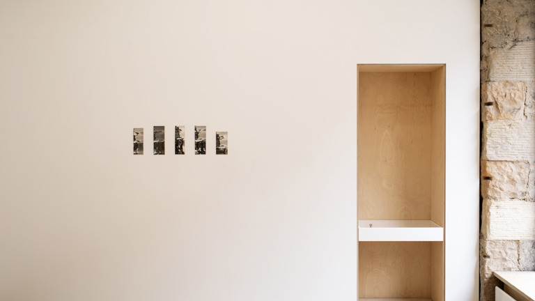 Minimalist gallery installation view showing a row of five small black-and-white photographs on a pale wall, beside a plywood-lined alcove with a built-in shelf and small object, exposed stonework visible to the right.