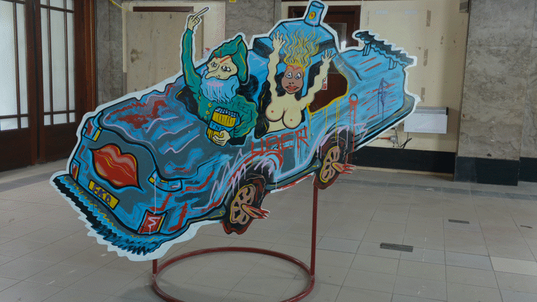 A plywood board held by a metal base has a drawn blue car with two people in it in Kelvin Hall as a part of Glasgow International 2018.