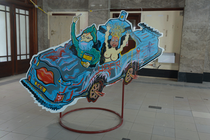 A plywood board held by a metal base has a drawn blue car with two people in it in Kelvin Hall as a part of Glasgow International 2018.