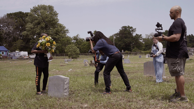 Colour photograph showing give individuals gathered around a small gravestone. One person holds a bouquet of flowers in front of their face, covering it, while the other four appear to be documenting the scene using cameras and audio equipment. 