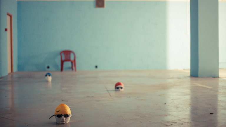 Le Swimming - sculptural heads in swimming goggles and caps laid out on the floor, as if in a swimming pool